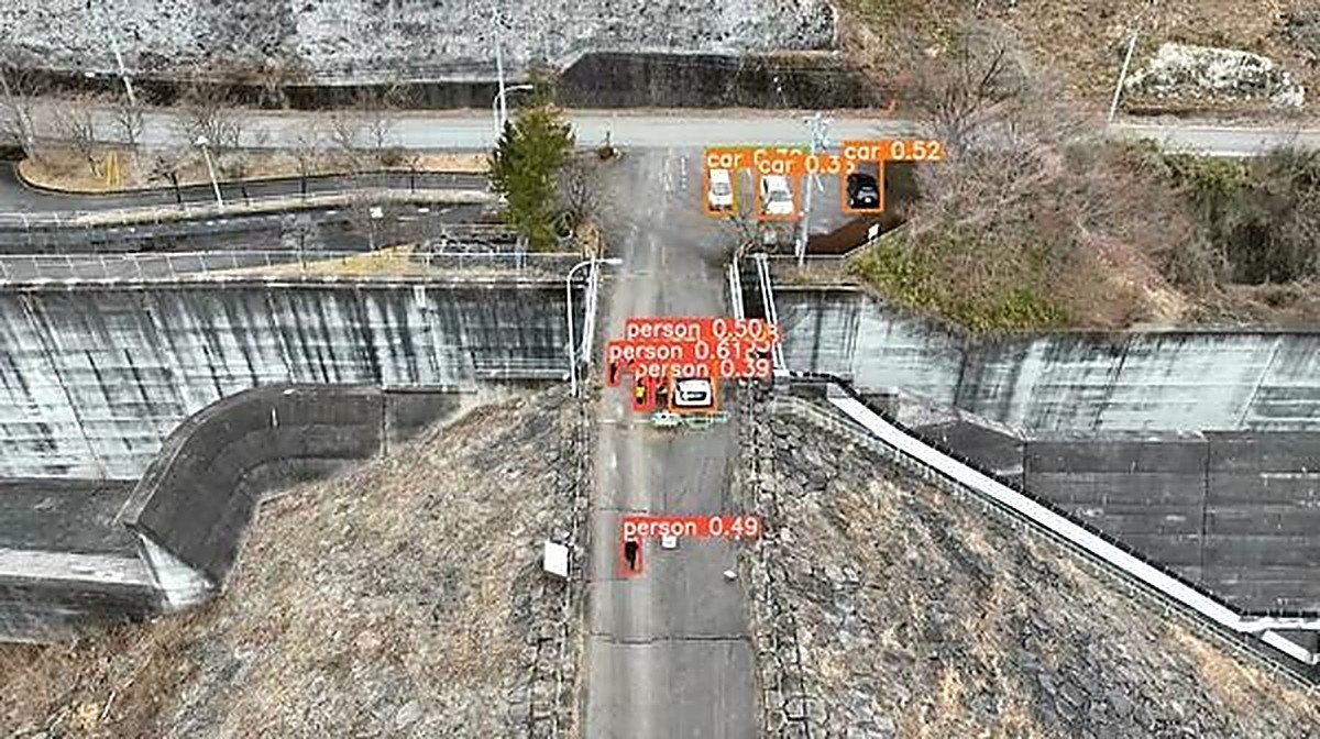 Aerial drone view of a fortified access road with live AI object-detection boxes over vehicles and pedestrians.